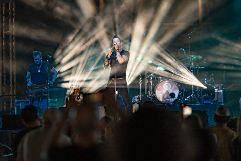 Scott Stapp sings at a microphone as beams of stage light shine over the concert crowd.