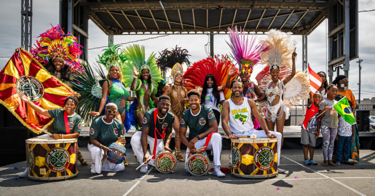 Unidos Atlanta Escola de Samba performers pose with drums and colorful feathered headdresses at the International City Experience.