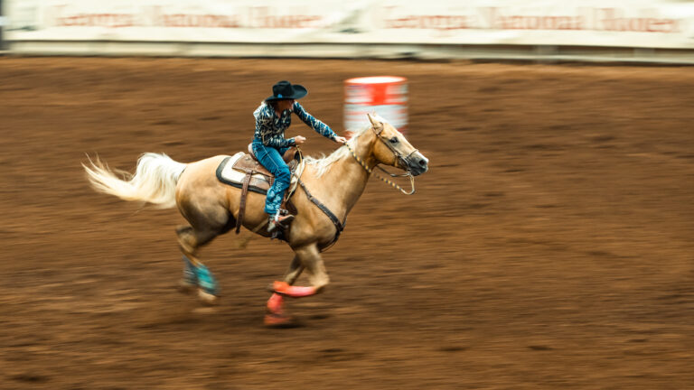 A barrel racer on a light-colored horse gallops across the indoor rodeo arena.
