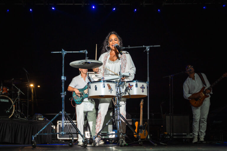 A singer performs behind a set of drums on a brightly lit outdoor concert stage at night.