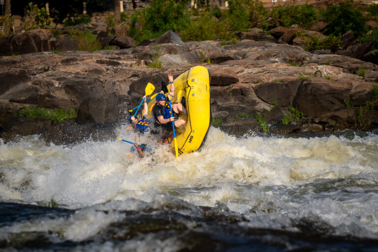Rafters in helmets and life jackets ride a yellow raft tipped upward in the Chattahoochee River rapids.
