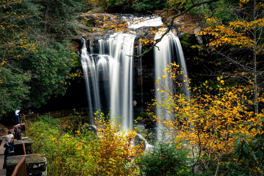Dry Falls waterfall cascading over rock with fall foliage and visitors on walkway behind the falls