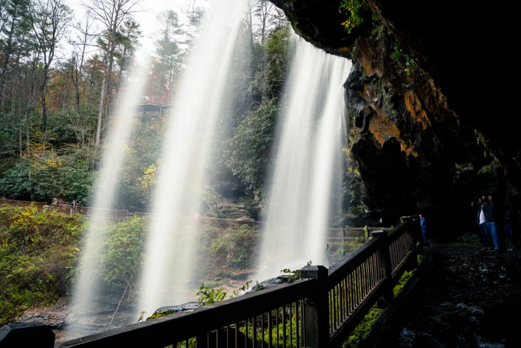 View from behind Dry Falls waterfall with railing and rock overhang in Highlands, North Carolina