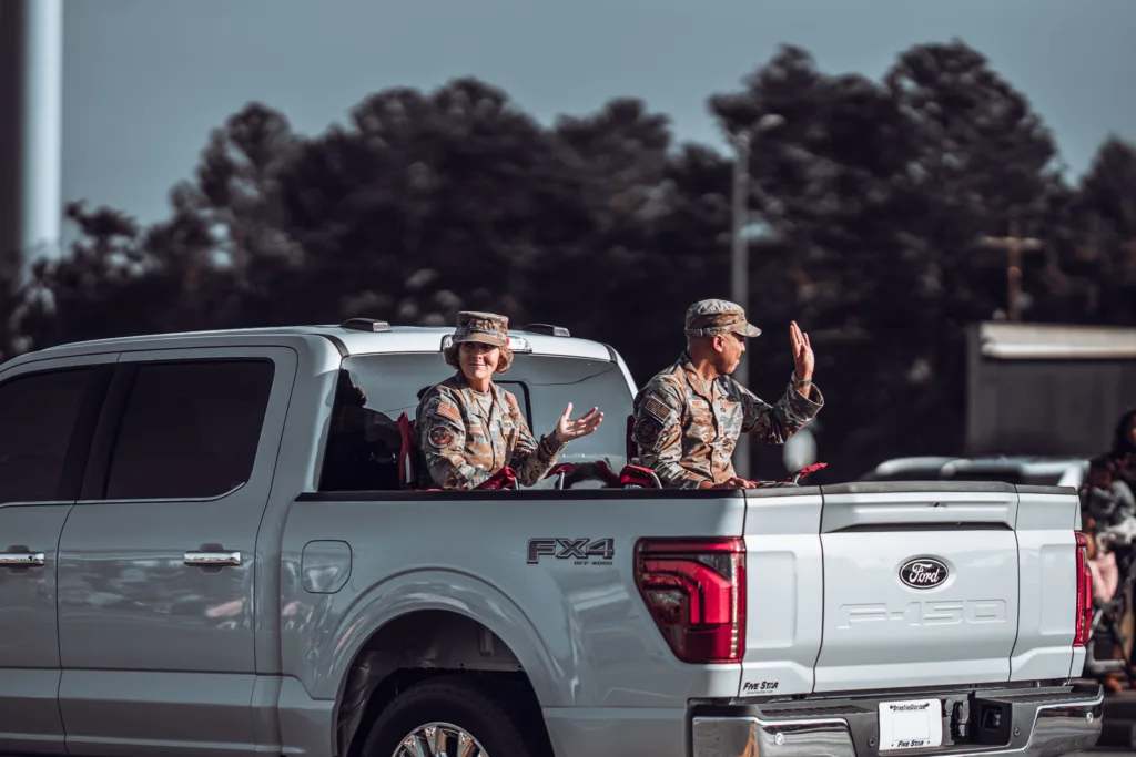 Two uniformed service members ride in the back of a silver pickup truck during a parade