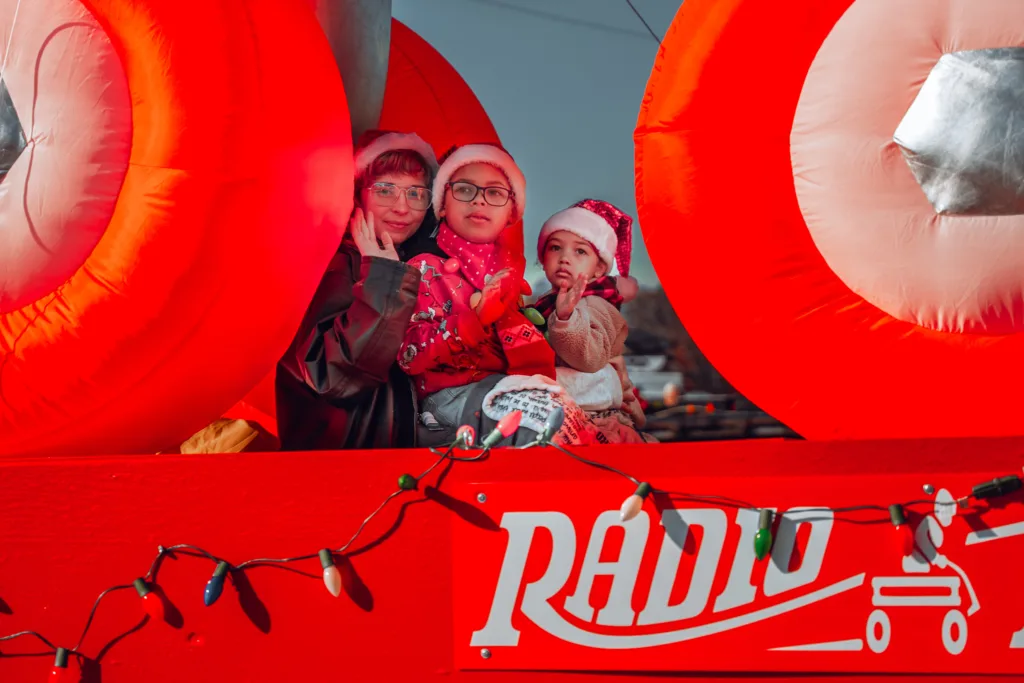 Three children in Santa hats riding a bright red holiday parade float