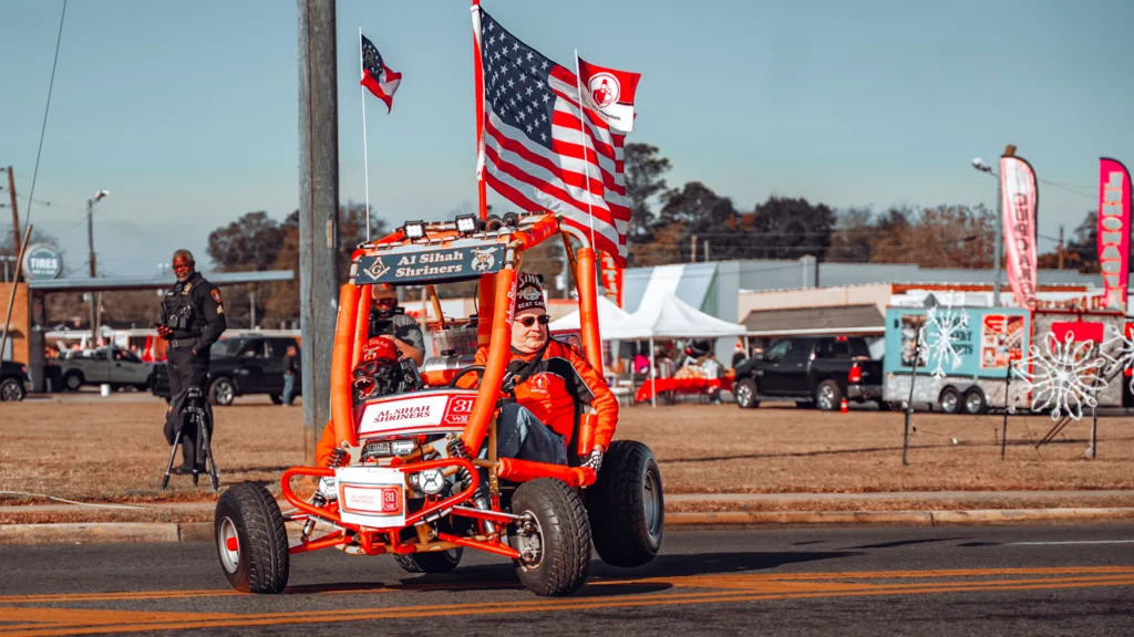 Shriners driving an orange parade car with American flag on a city street
