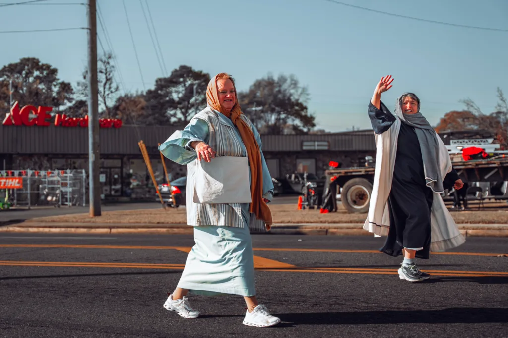 Two women in robes and headscarves walking and waving in a street parade