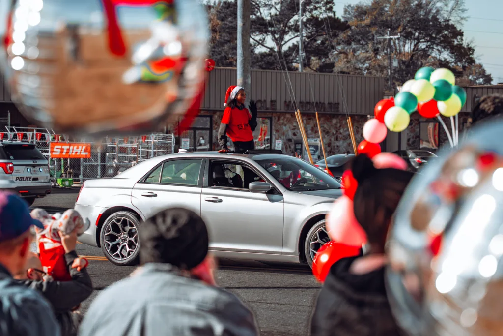 Silver car in a parade with person standing through sunroof and balloons