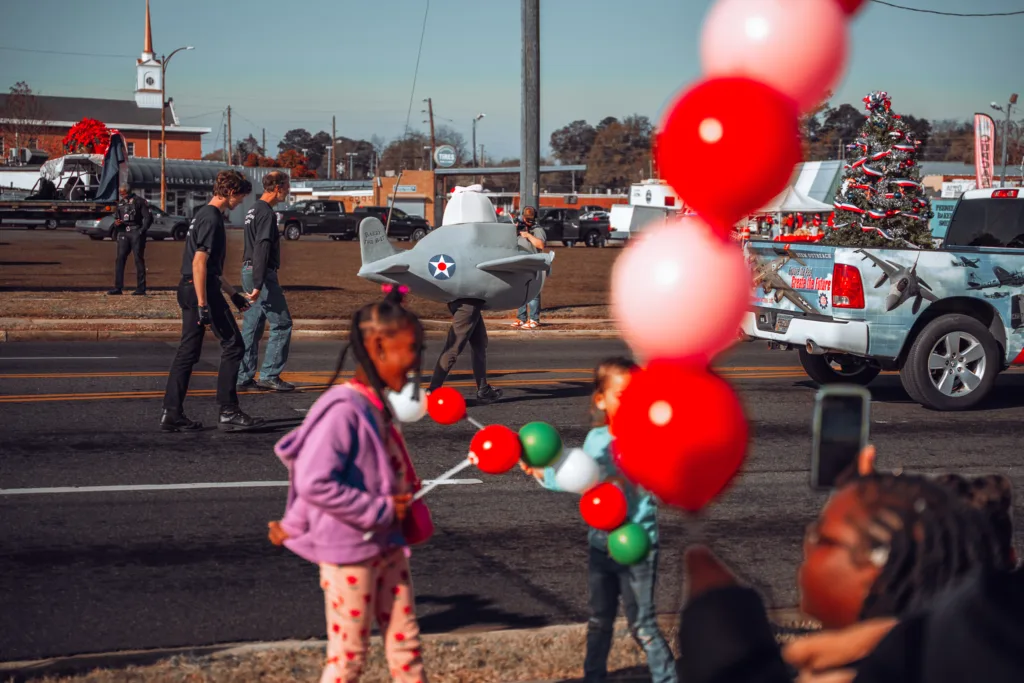Children holding colorful balloons while a holiday parade passes by on a street