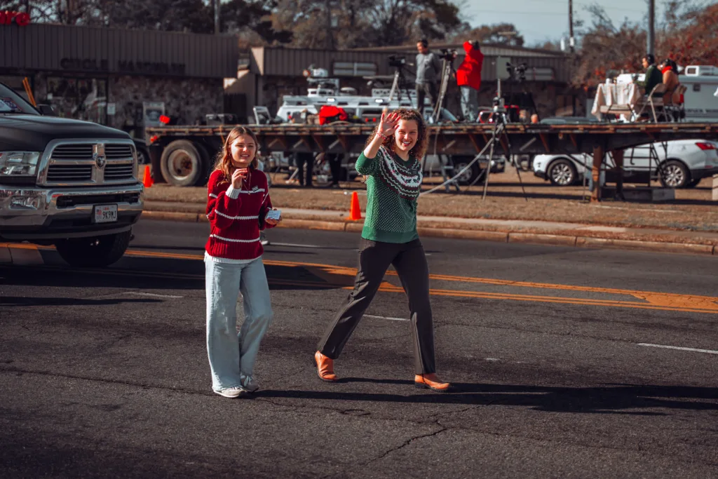 Two people in sweaters wave while walking down a city street parade route