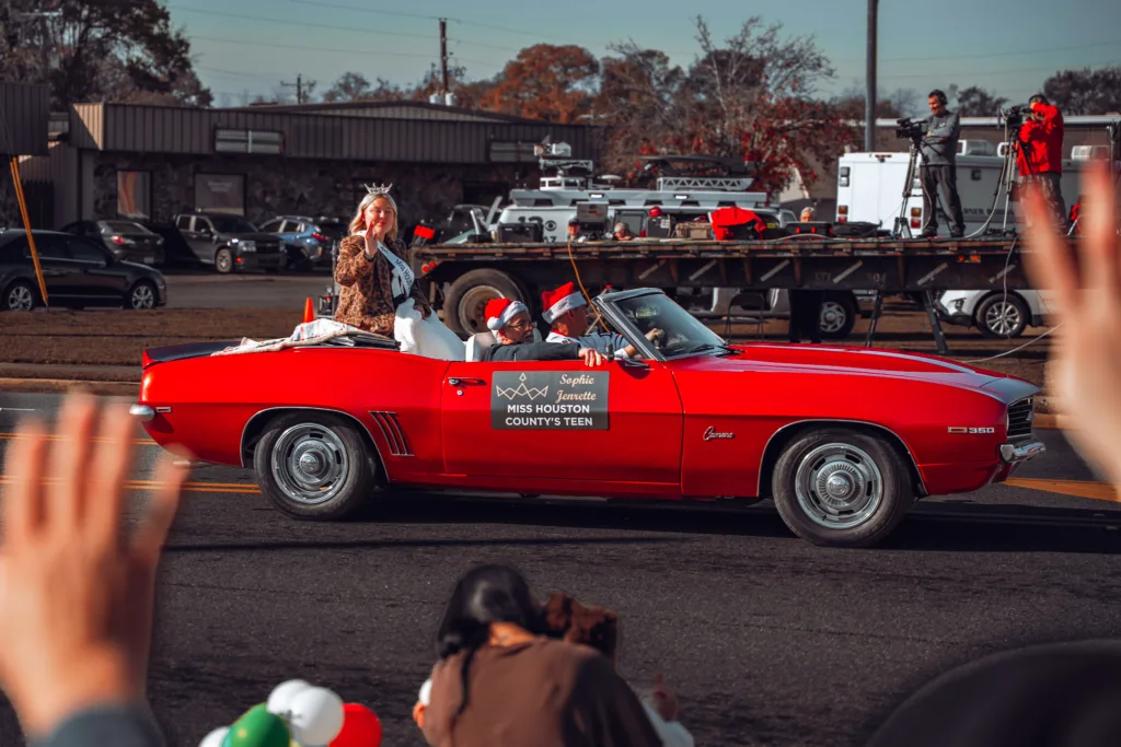 Woman waves from a red convertible during a parade as spectators watch from the roadside