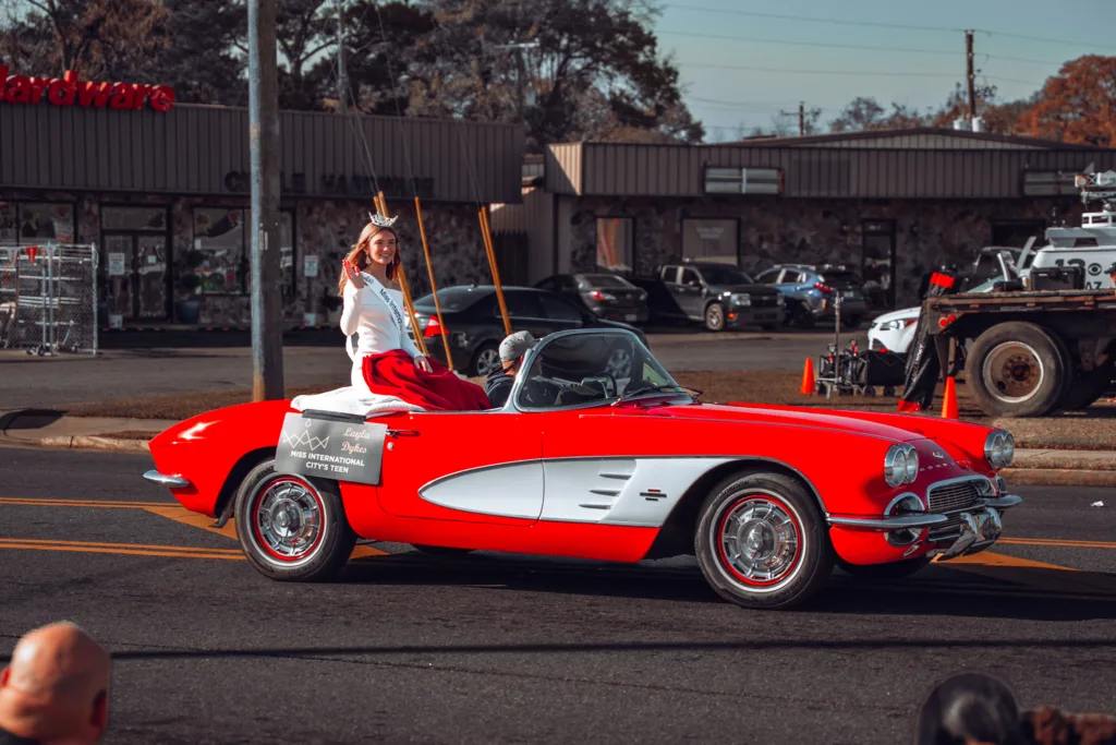 Woman waves from a red vintage convertible driving in a street parade with storefronts and spectators in the background