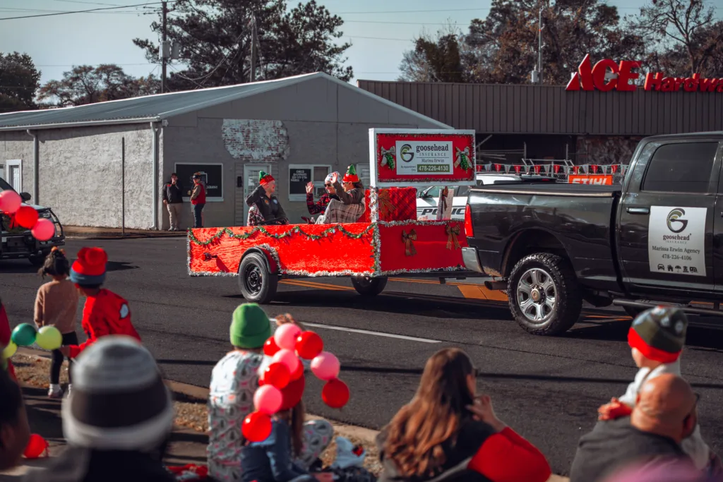 Holiday parade float with red garland towed by a pickup truck as spectators watch along a small-town street