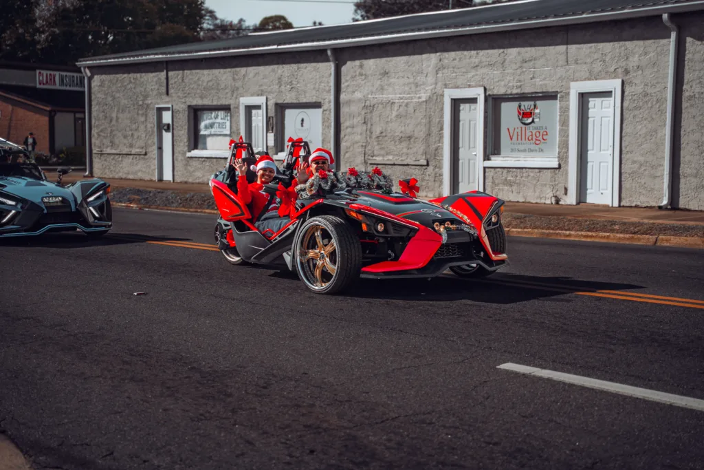 Red Polaris Slingshot three-wheeler with festive costumed riders parked on a small-town street in a parade lineup