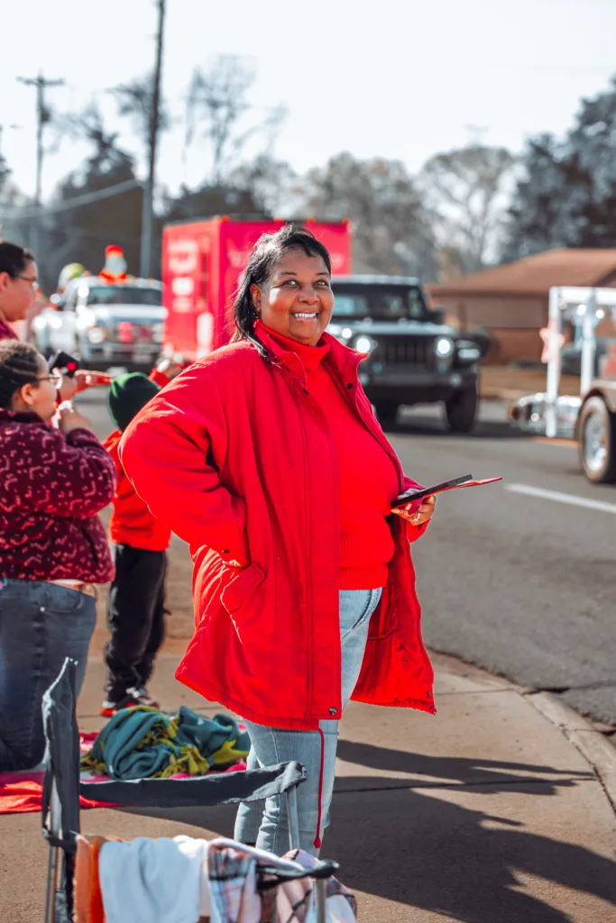 Smiling woman in a red coat holding a phone on a sidewalk as parade vehicles pass by on a sunny day