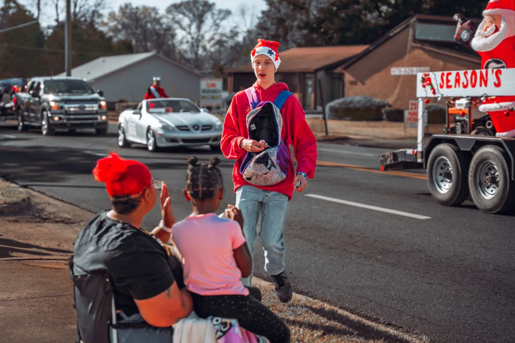 Holiday parade participant in a Santa hat and red hoodie walks past seated spectators as decorated vehicles drive by