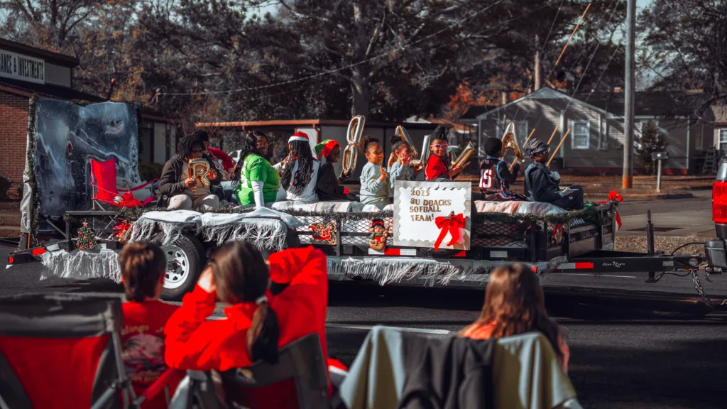Holiday parade float with people in Santa hats and festive decorations as spectators watch from folding chairs