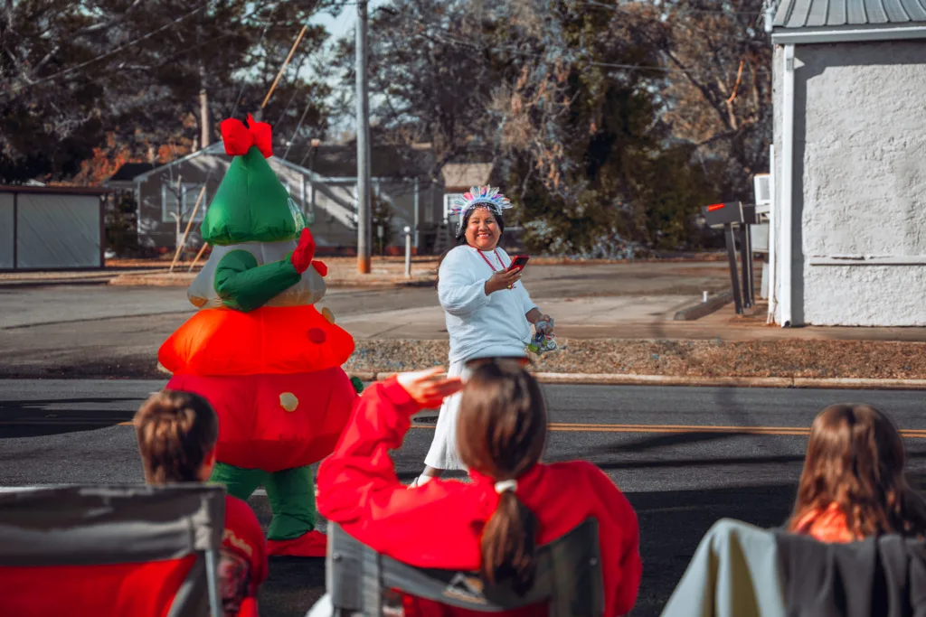 Holiday parade participant waves on a street beside a Christmas tree mascot as spectators watch from folding chairs