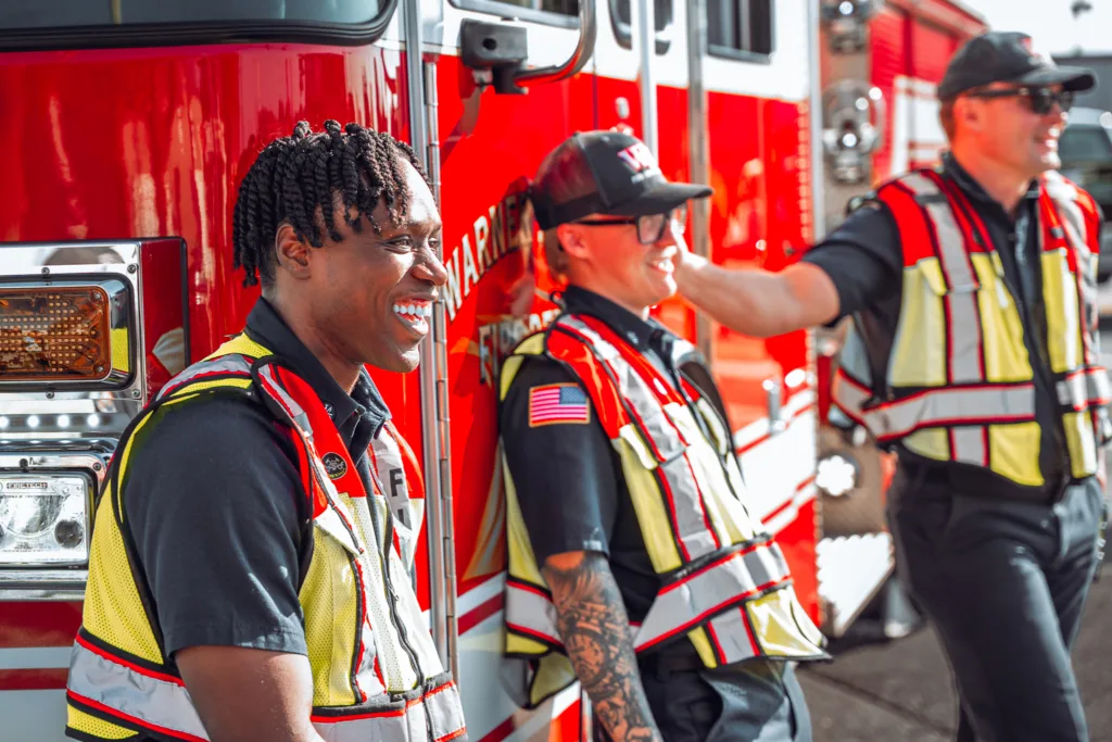 Firefighters wearing reflective safety vests stand beside a red fire truck in bright daylight at an outdoor event.