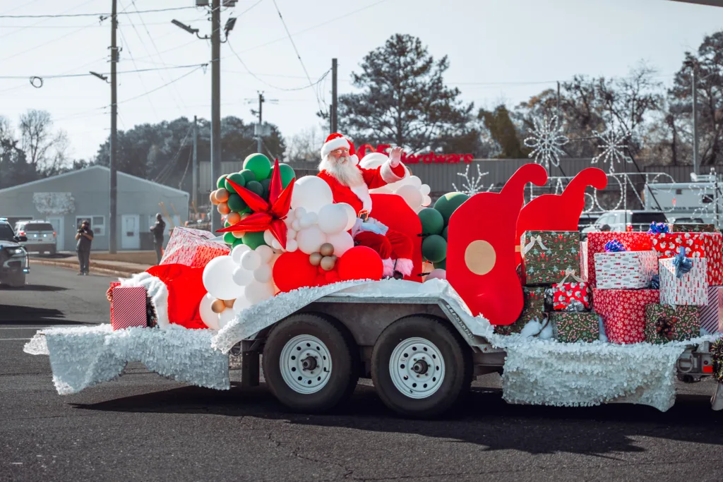 Santa Claus on a holiday parade float with a red sleigh, balloons, and stacks of wrapped Christmas presents on a street