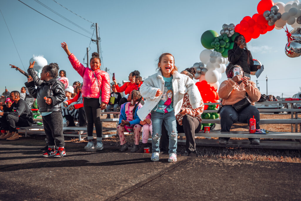 Smiling children cheer and dance in front of bleachers at a parade