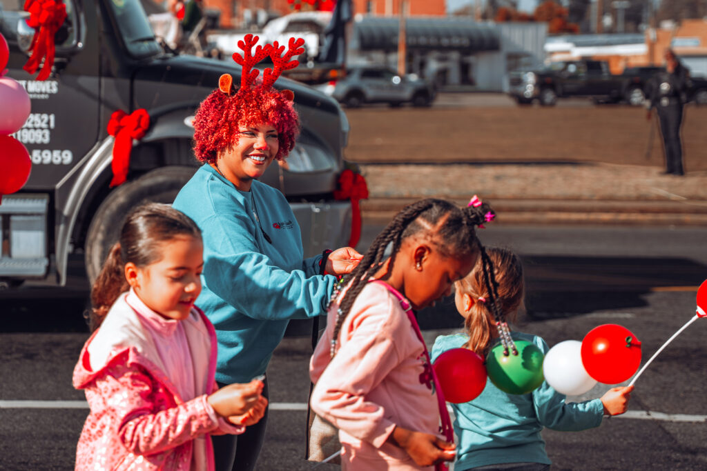 Smiling woman with reindeer antlers and children holding balloons at holiday parade
