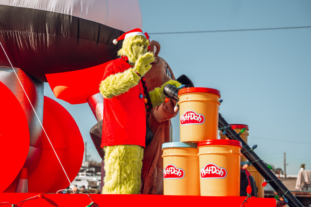 Grinch character in red shirt standing on bright parade float with Play-Doh props