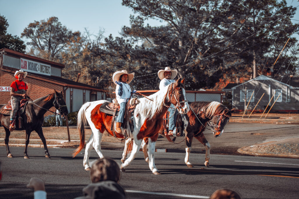 Cowboys in hats ride horses along a street during a small-town parade as spectators watch from the roadside