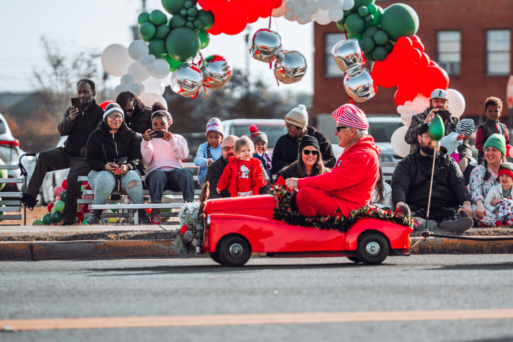 Man in red suit drives tiny red car past families at holiday parade