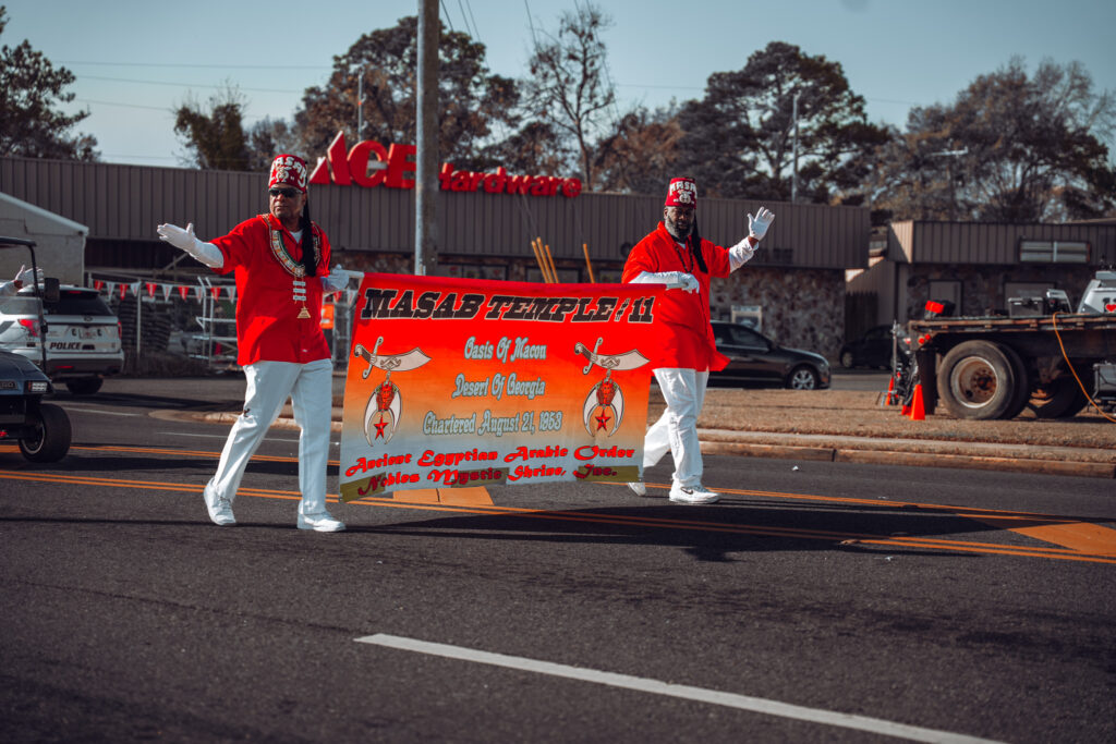 Two parade participants in red outfits carrying a Masab Temple banner on a city street