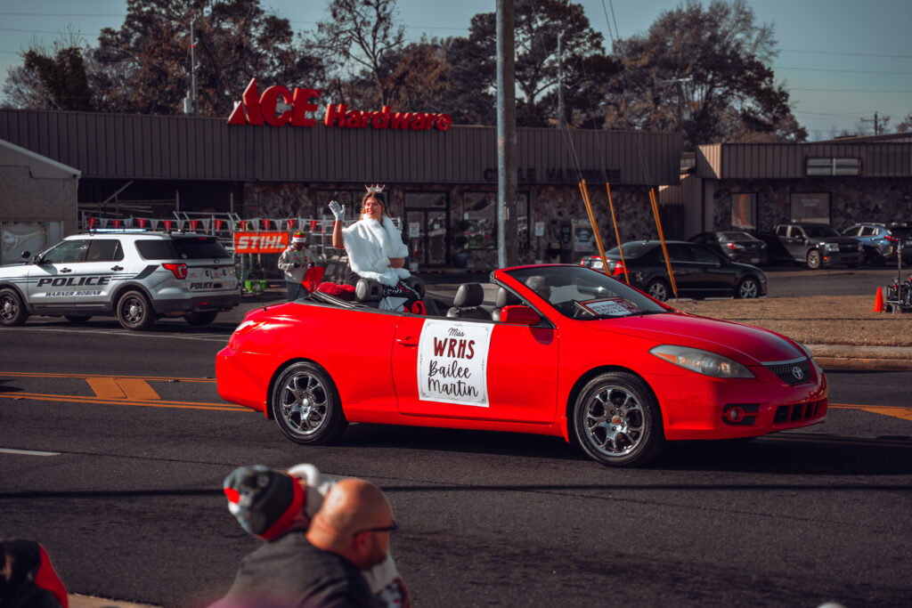 Person waving from a red convertible in a parade on a city street, with police SUVs and storefronts in the background