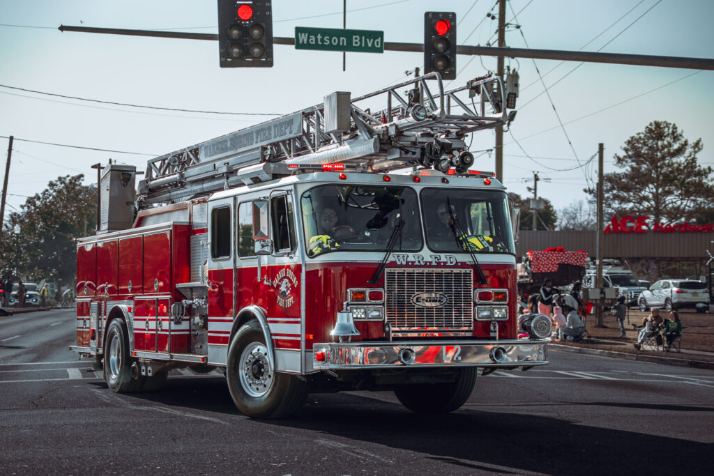 Red ladder fire truck driving through a city intersection under a Watson Blvd street sign with traffic lights, cars, and powe