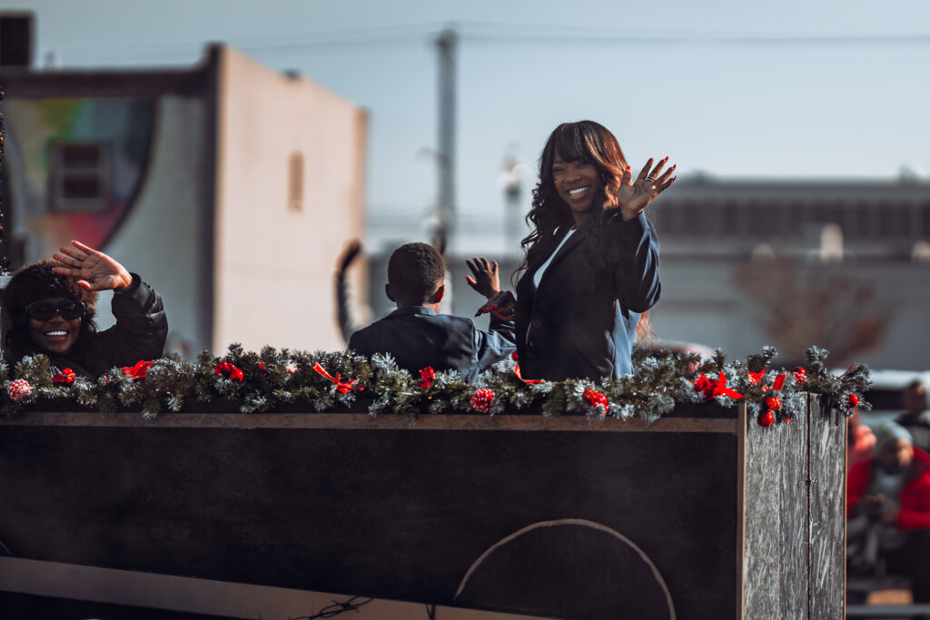 Smiling woman waves from a garland-decorated parade float during a holiday event