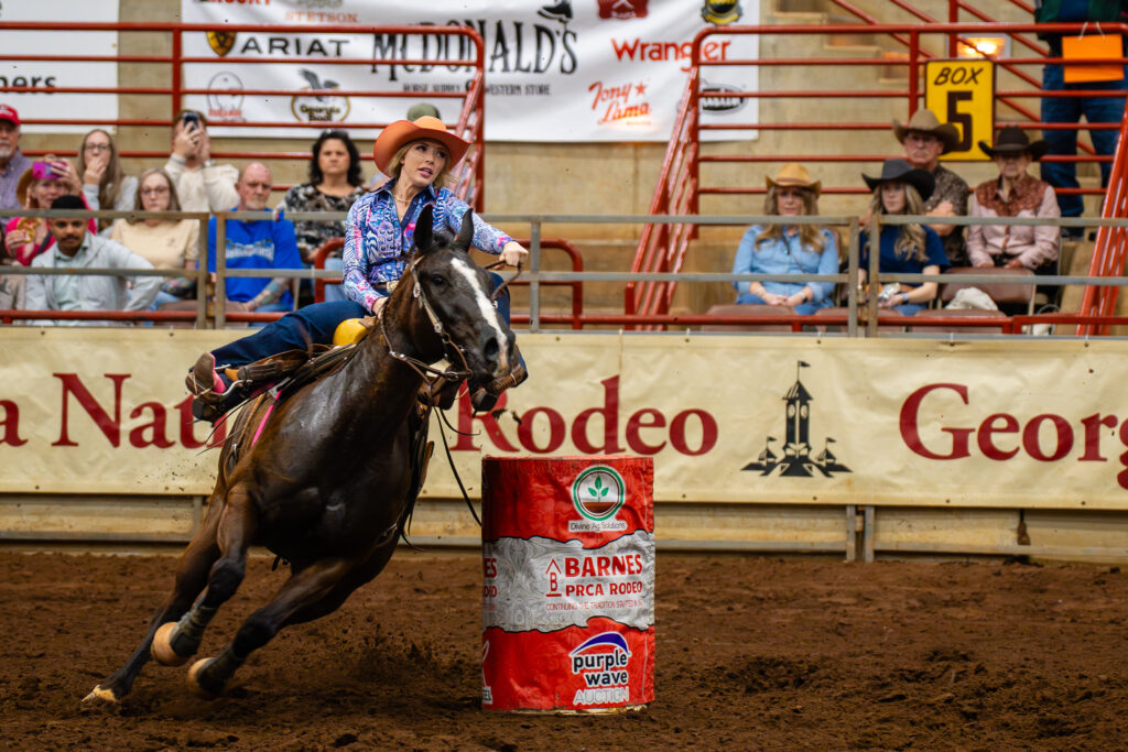 Cowgirl on galloping horse rounding a barrel in an indoor rodeo arena