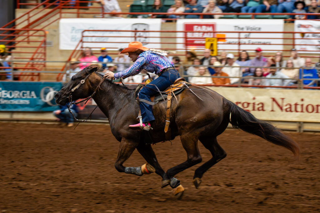 Barrel racer on galloping horse in indoor rodeo arena with crowd