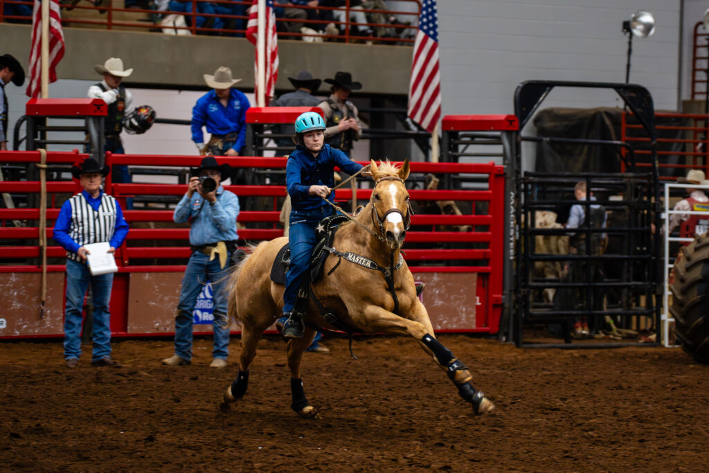 Young rider in helmet racing a palomino horse around a barrel in an indoor rodeo arena