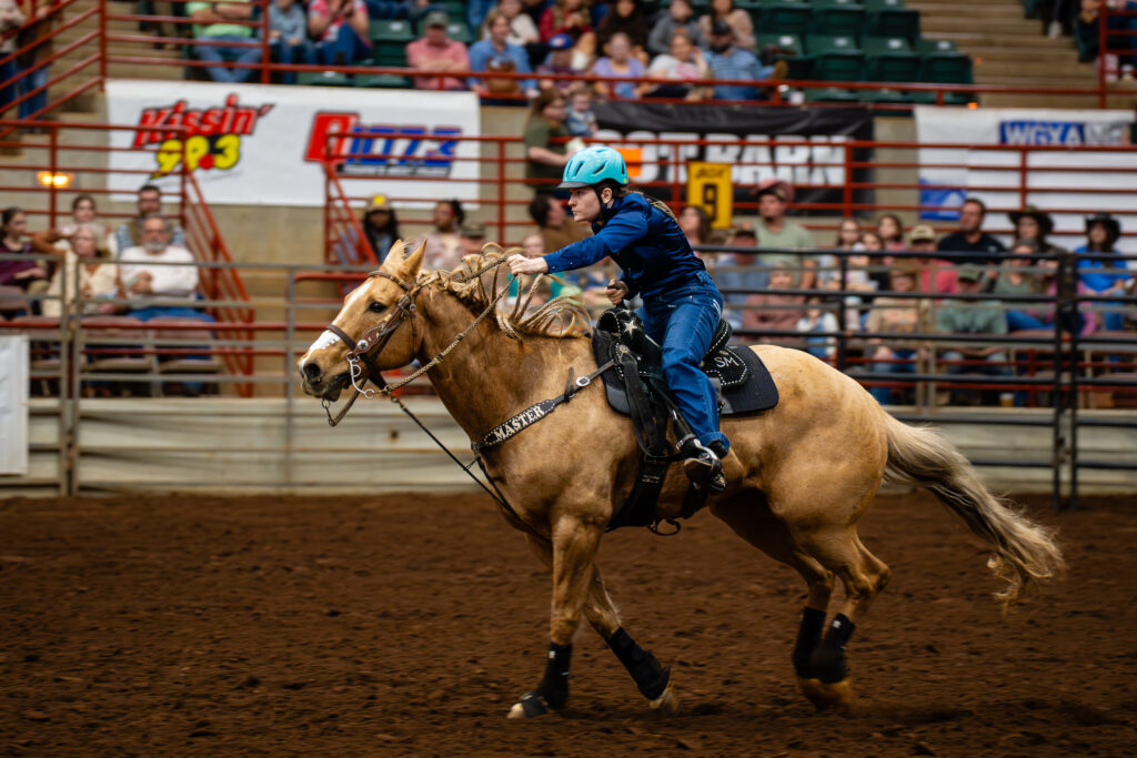 Barrel racer in blue helmet riding a palomino horse in an indoor arena