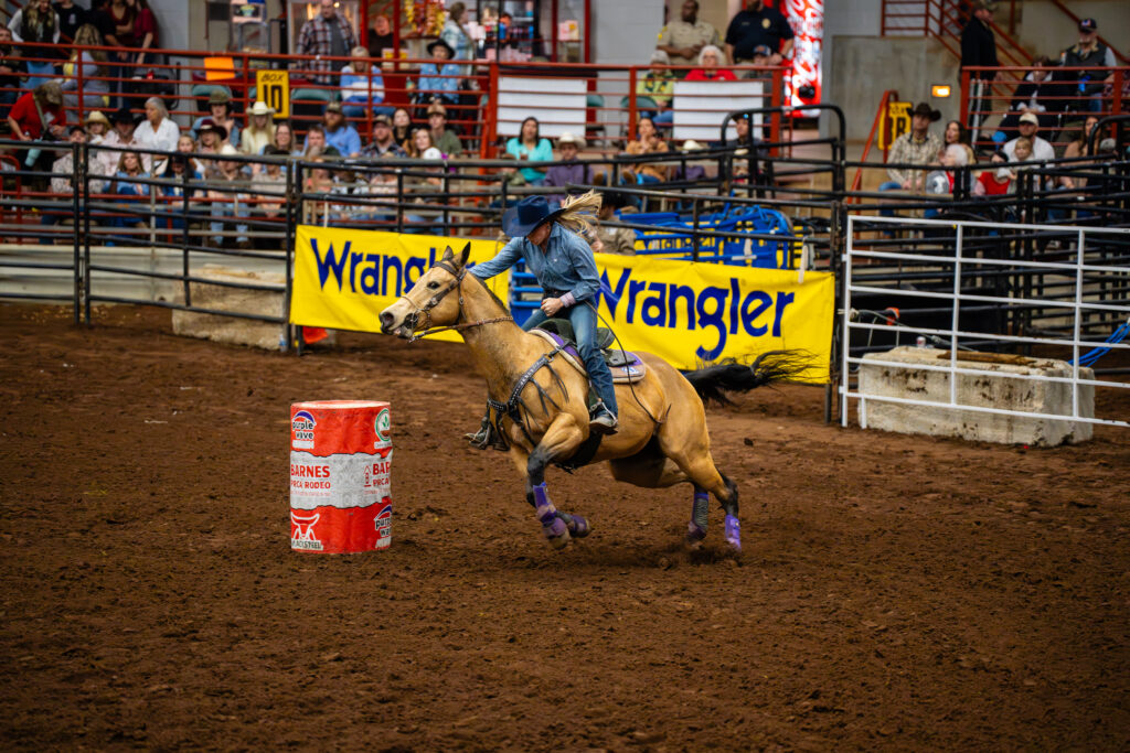 Cowgirl barrel racing on a horse rounding a barrel in an indoor rodeo arena