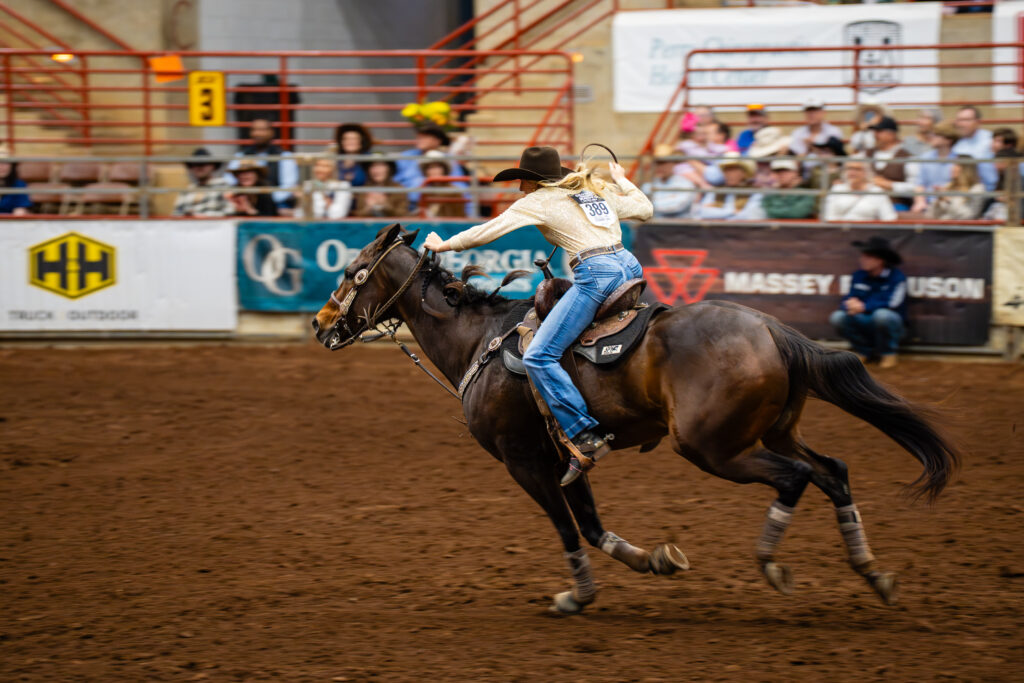 Barrel racer in cowboy hat galloping a horse across an indoor rodeo arena