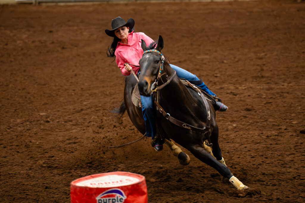 Cowgirl in pink shirt barrel racing on a dark horse in a dirt arena