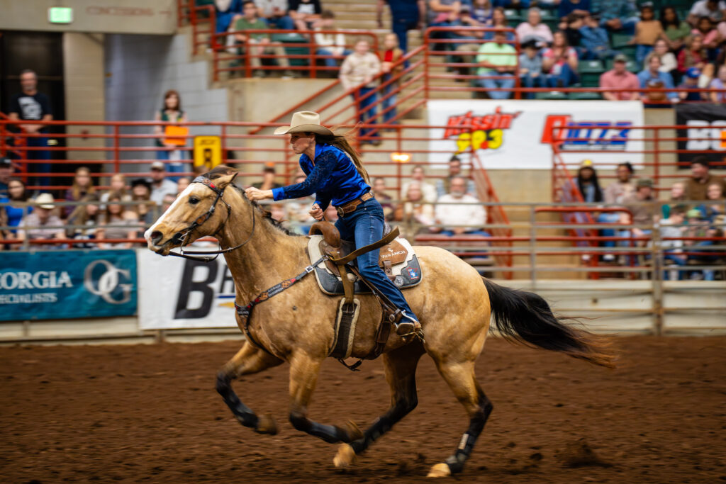 Cowgirl in blue shirt barrel racing a tan horse across an indoor rodeo arena