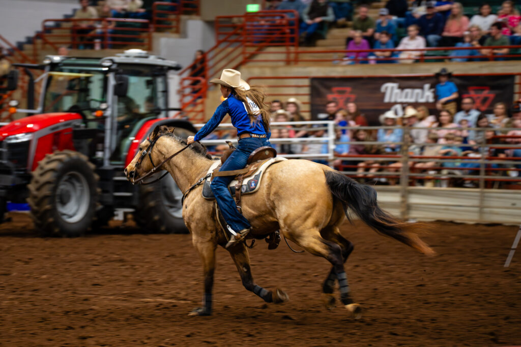 Cowgirl in blue shirt barrel racing on tan horse in indoor rodeo arena