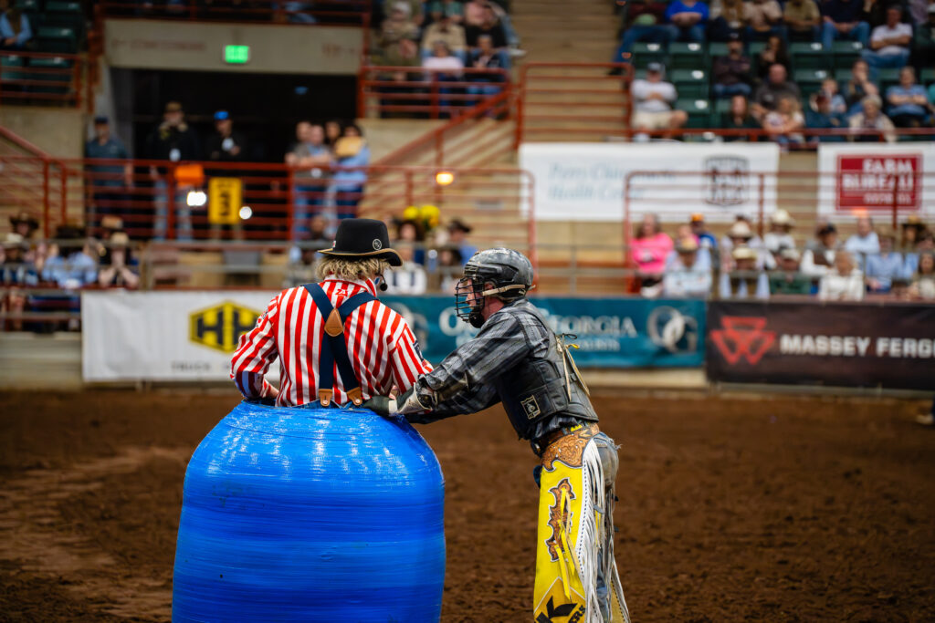 Bull rider in safety gear talks with rodeo clown beside blue barrel