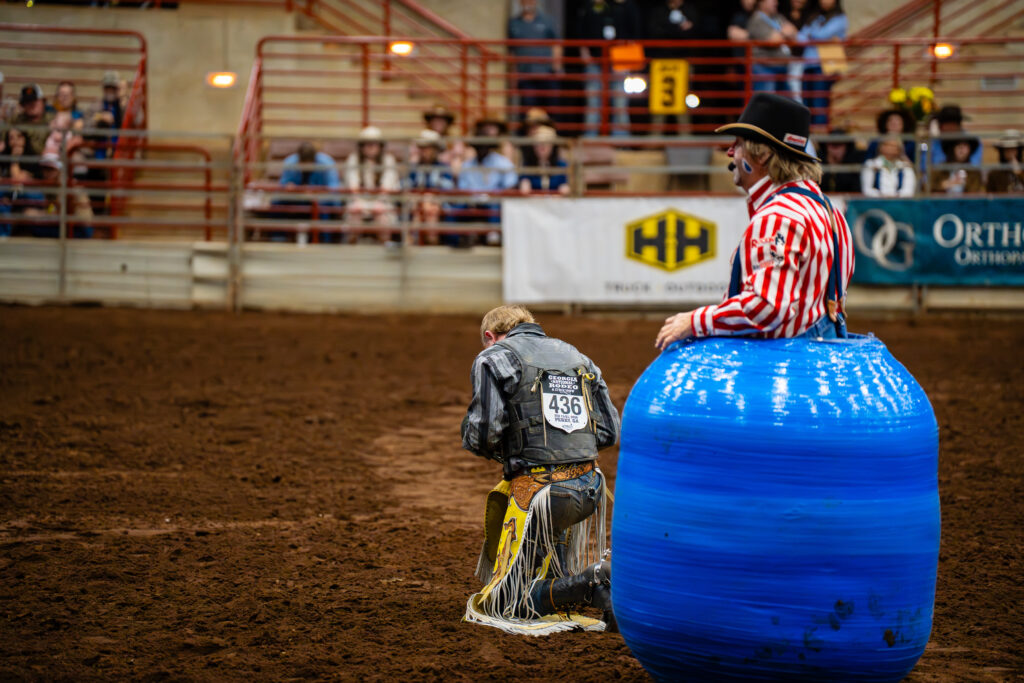 Bull rider kneeling in dirt arena beside rodeo clown in blue barrel