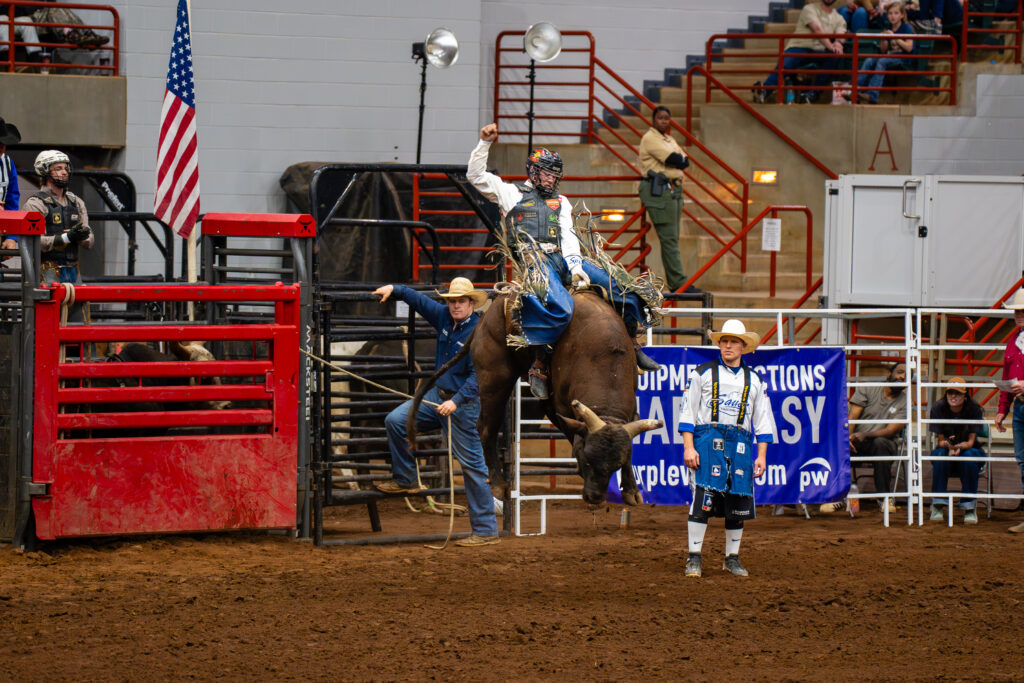 Bull rider holding one arm up while riding a bucking bull in an indoor rodeo arena