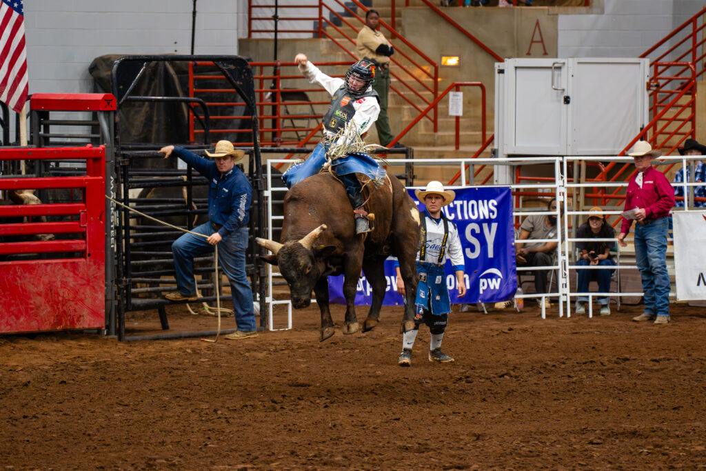 Bull rider holding one arm up while riding a bucking bull in an indoor rodeo arena