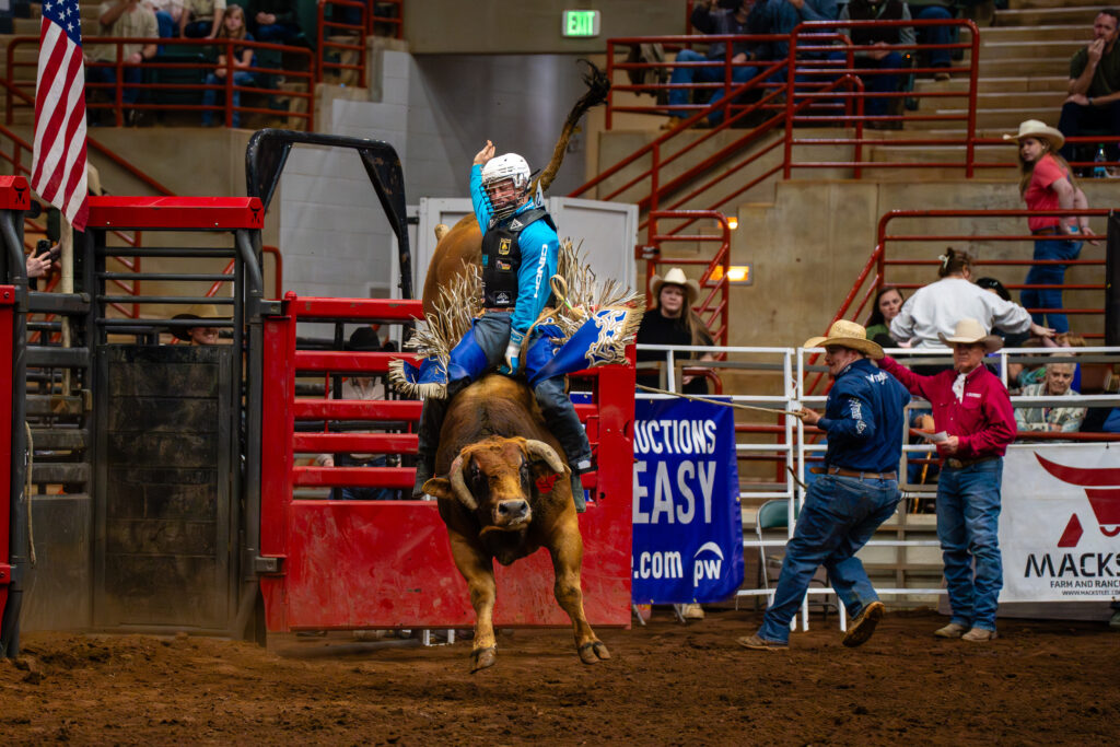 Bull rider in blue gear holding on during ride at indoor rodeo