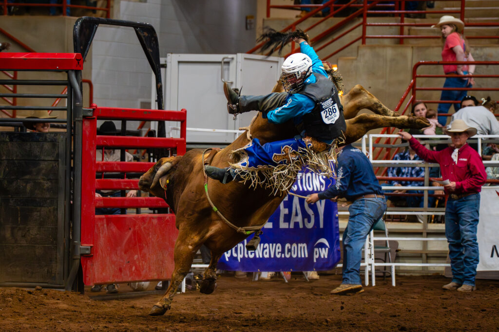 Bull rider in blue gear thrown into the air by a bucking bull at a rodeo arena