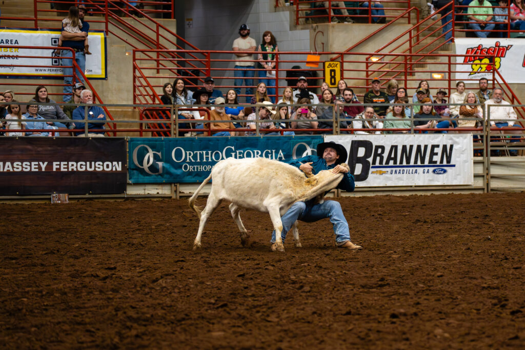 Cowboy wrestling a steer to the ground in a crowded indoor rodeo arena