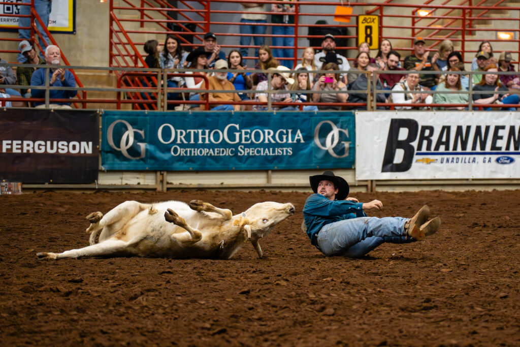 Steer wrestler sliding in dirt beside calf during indoor rodeo event